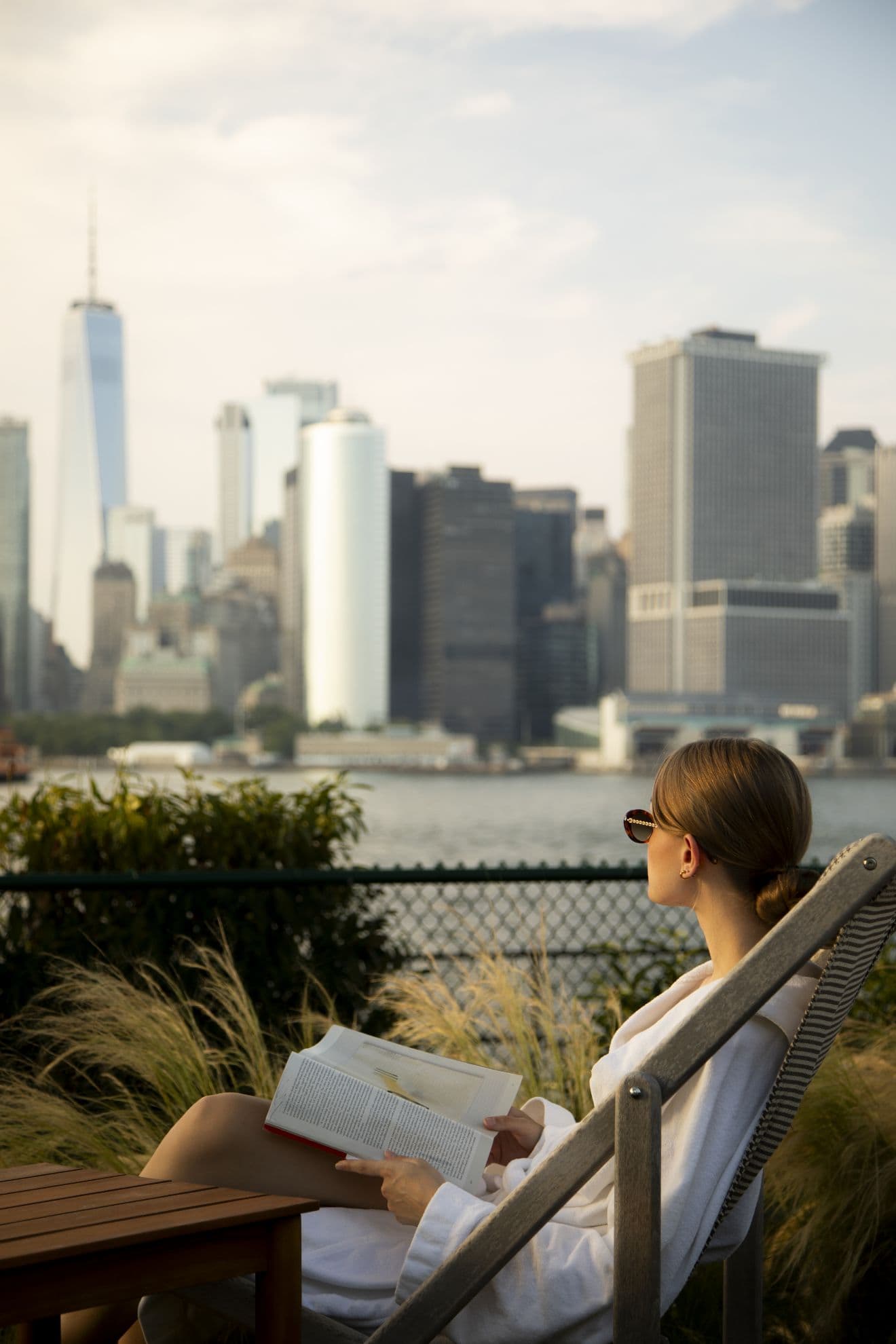 A woman wearing sunglasses, sitting on a deck chair with a book in her hand, looking out at the New York skyline from the garden